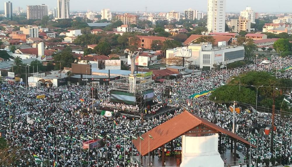 Landscape distance photo of large amount of people gathered near wooden structure in the foreground with a city scape in the background. In the middle, there is a structure displaying a green and white striped flag